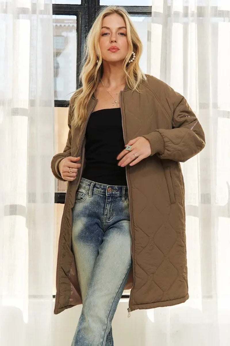 woman wearing brown zip up long utility jacket with pockets and quilted design over black top and blue jeans in bright room