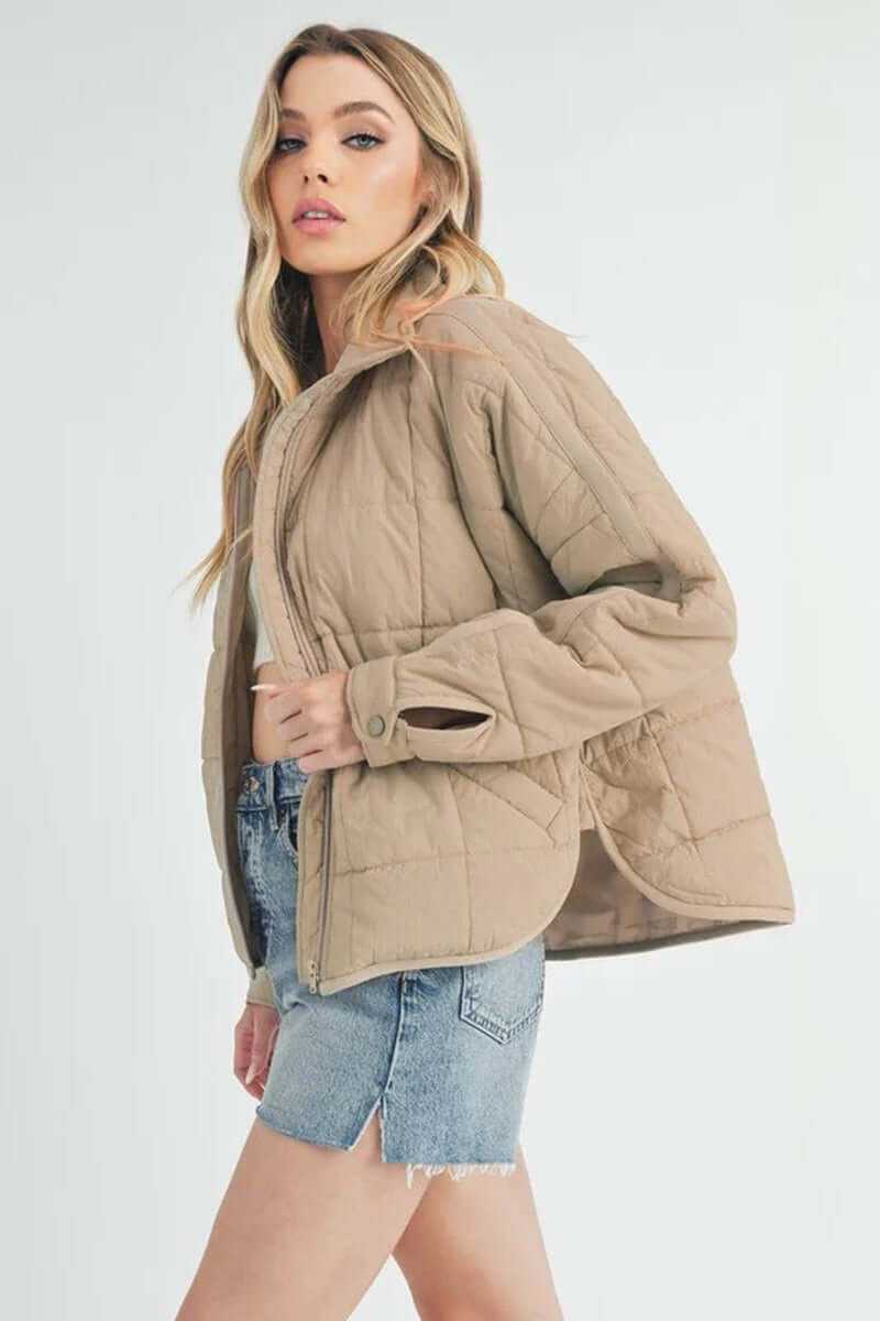 Bebe puffer jacket in beige with oversized fit, front zip closure, and snap button cuffs, styled with denim shorts.