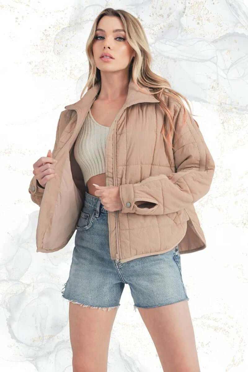 Model showcasing Bebe puffer jacket in beige, styled with a cropped top and denim shorts against a marble background.