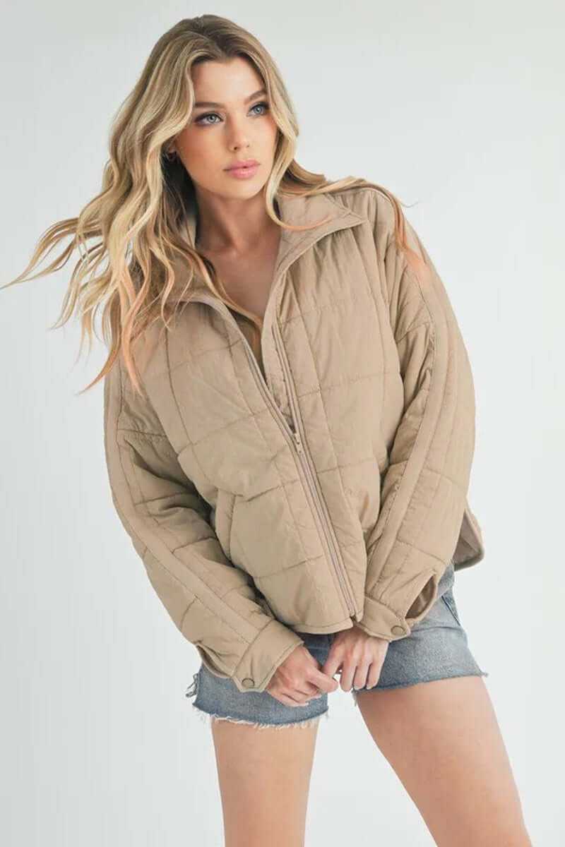 Female model wearing a beige Bebe puffer jacket with oversized fit and zip closure, styled with denim shorts.