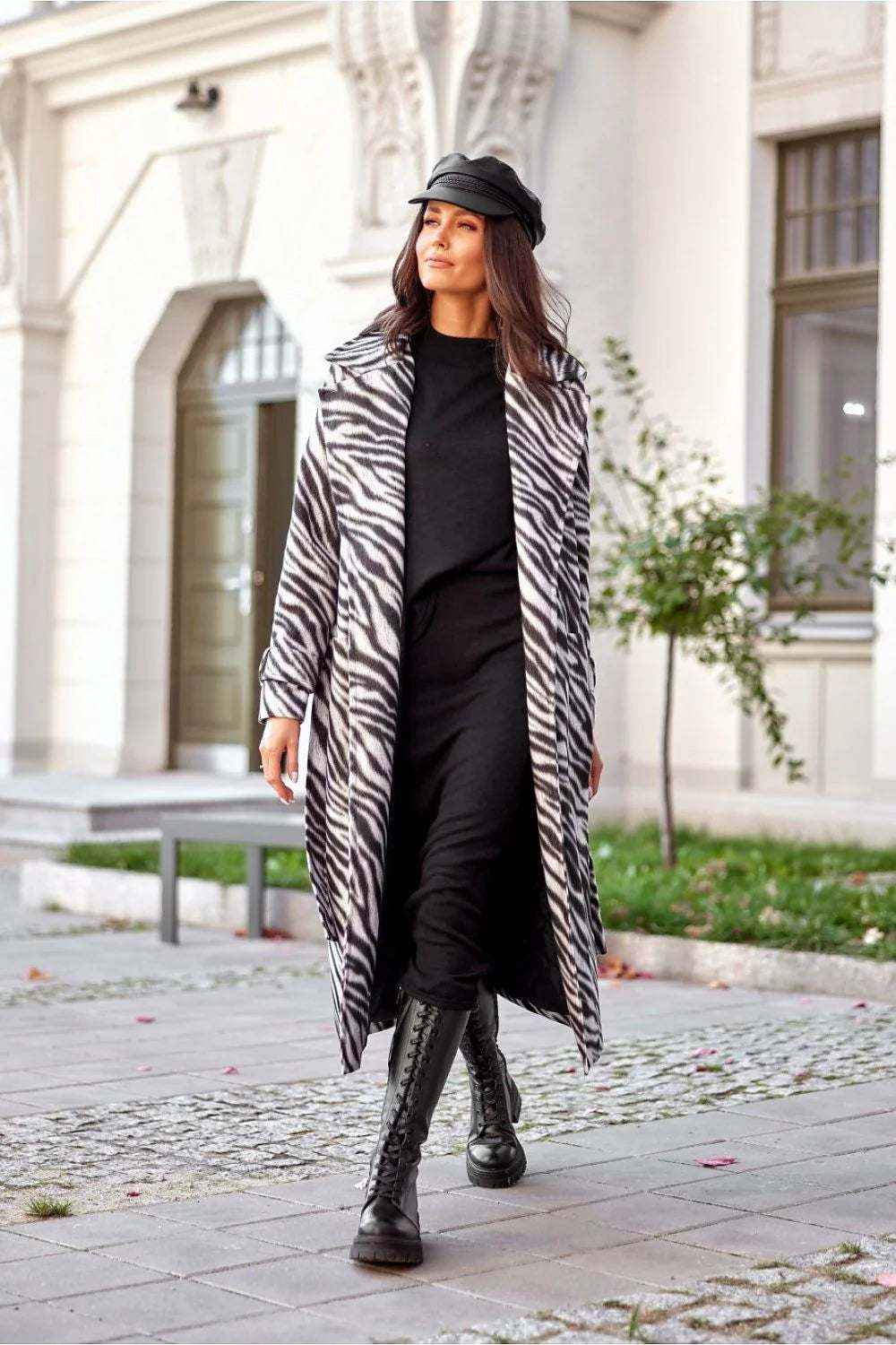Stylish woman in a zebra print longline coat walking outdoors with black boots and cap.