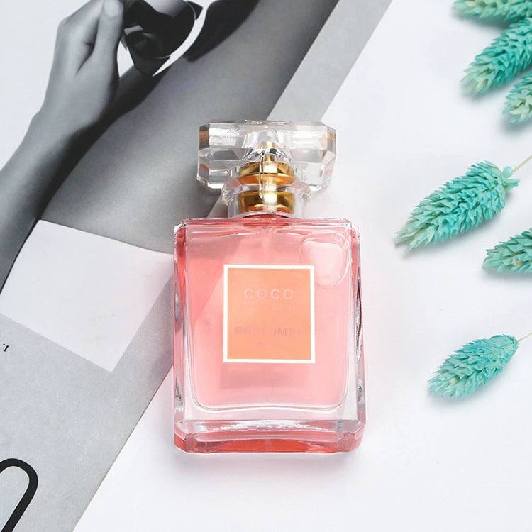 Clear glass perfume bottle with pink liquid and square acrylic cap on white background with decorative plants and magazine page