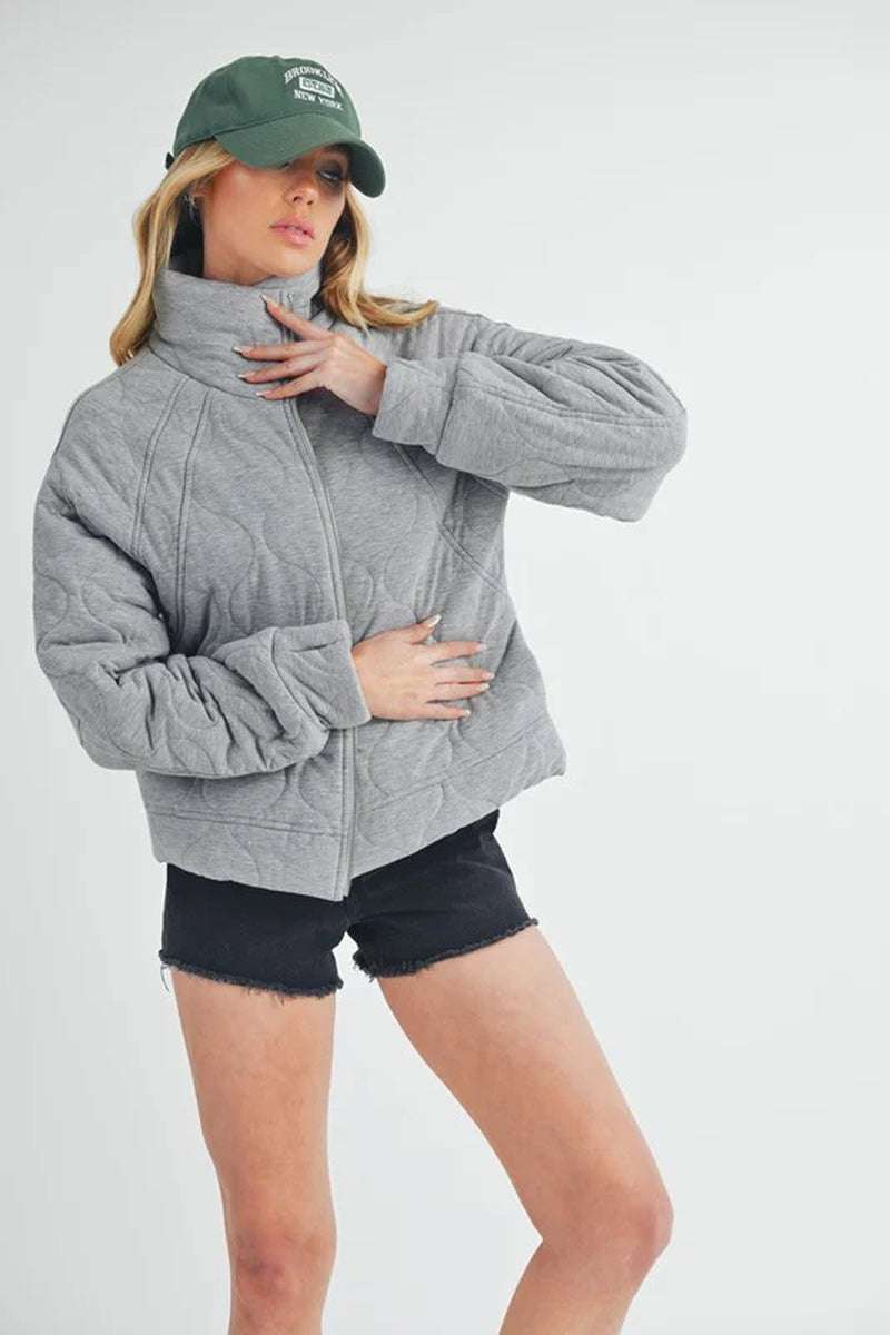 Woman wearing a gray Darla quilted jacket with oversized fit and front zipper, styled with black shorts and a green cap.
