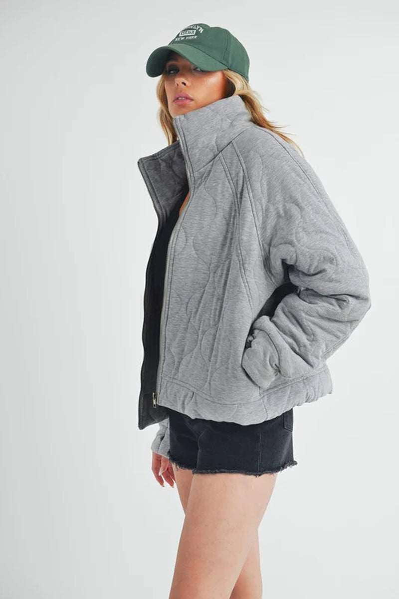 Woman wearing a cozy Darla Quilted Jacket in grey with a front zipper, stylishly paired with a green cap.