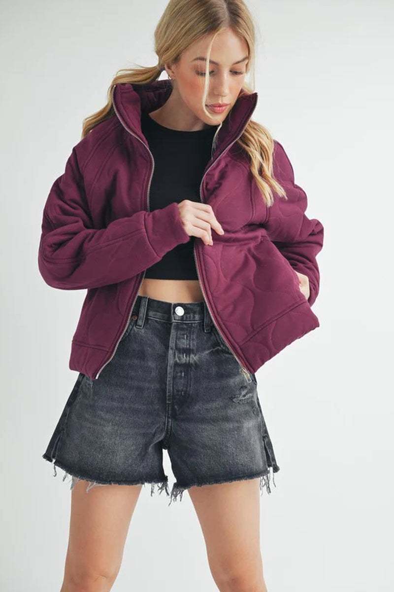 A woman models a cozy oversized burgundy quilted jacket paired with black denim shorts, showcasing a stylish fall outfit.