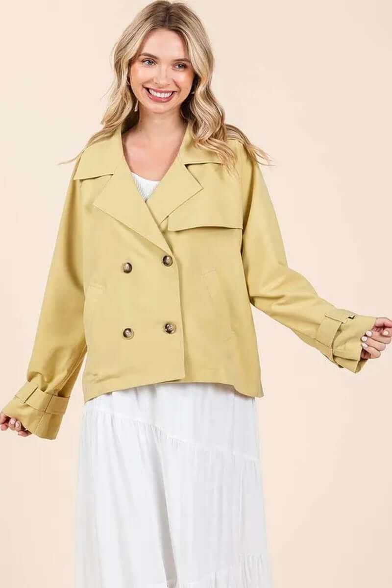 Model wearing a yellow double-breasted long sleeve trench coat jacket over a white dress, showcasing stylish outerwear.