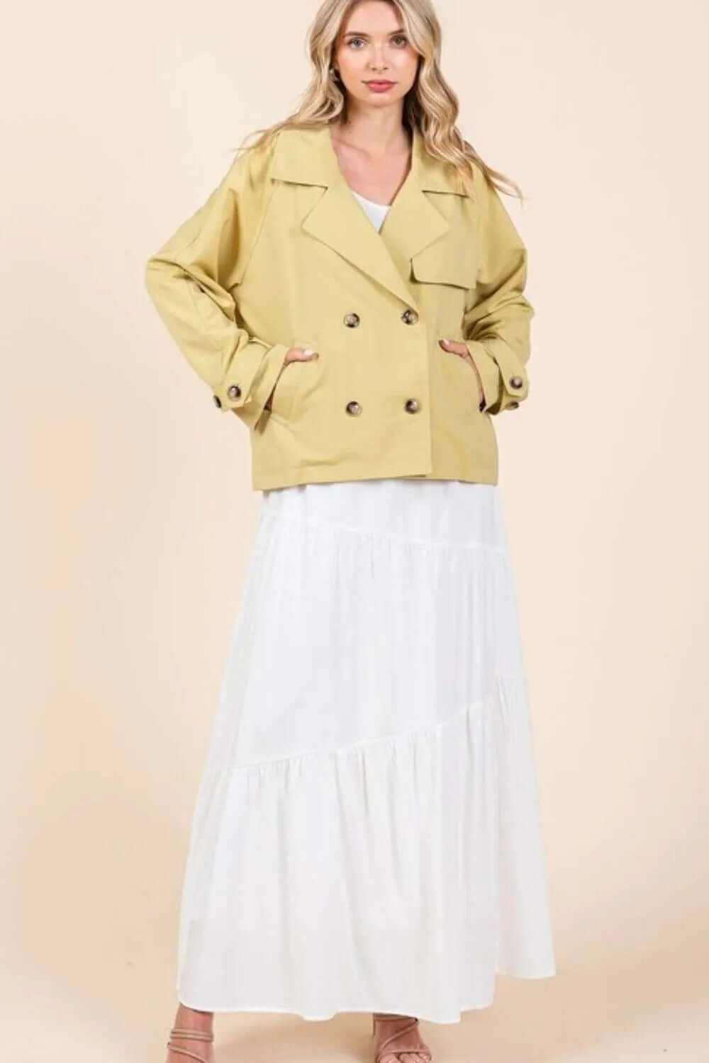 Woman wearing a double-breasted long sleeve trench coat jacket in mustard color over a white dress.