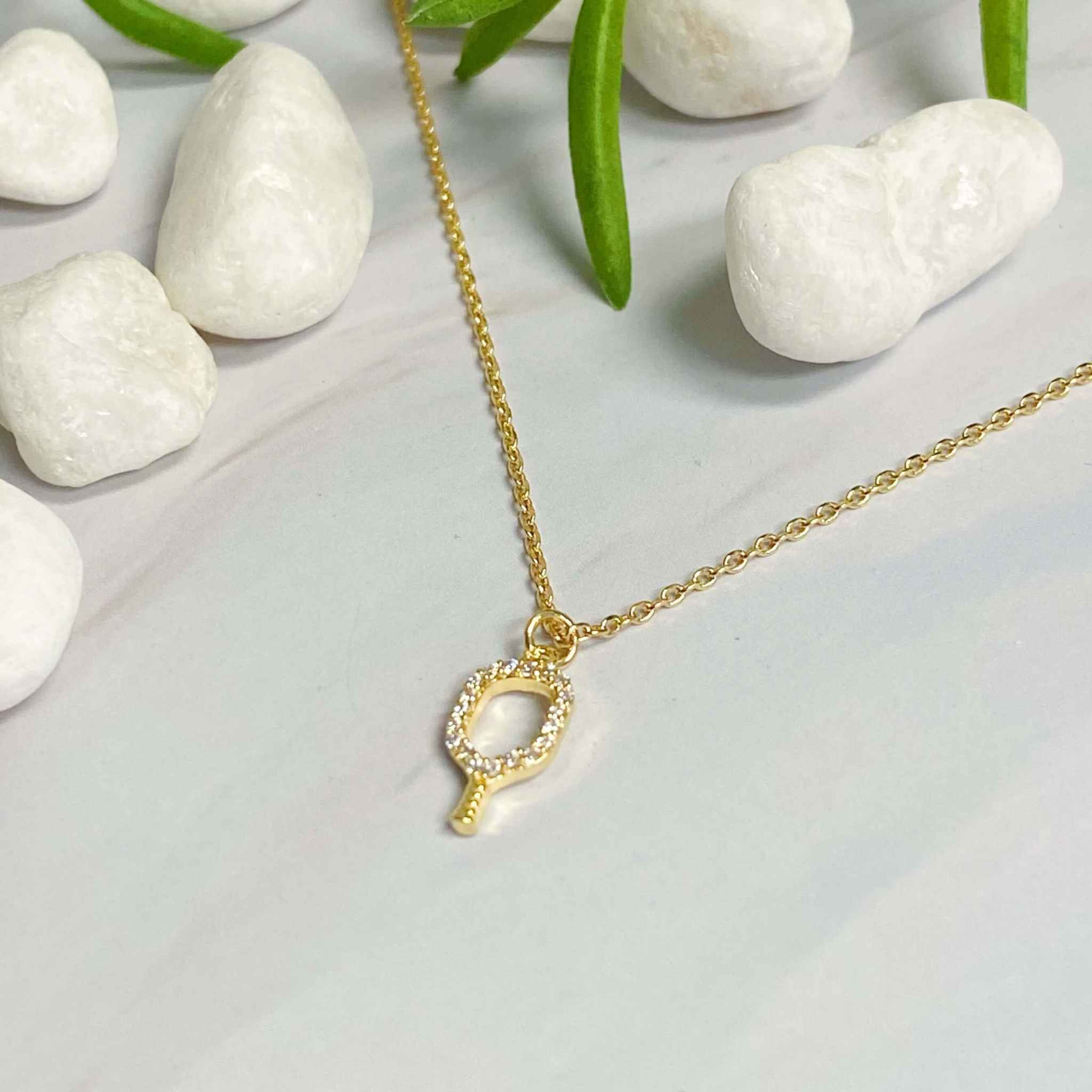 Dainty gold pickleball racket pendant necklace on a minimalist background with white stones.