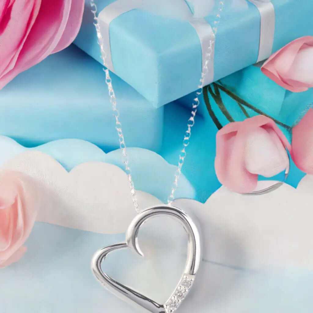 14K silver plated love heart pave pendant necklace next to pink flowers and a blue gift box.