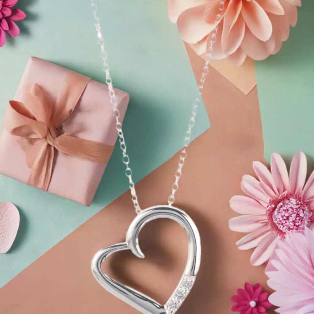 14K silver plated heart pendant necklace with cubic zirconia against a backdrop of flowers and a gift box.