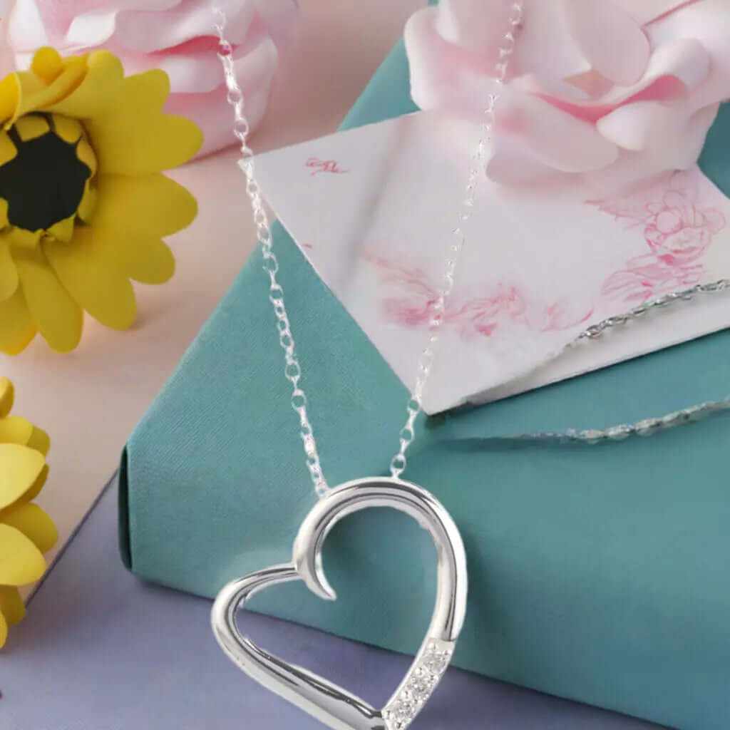 14K silver plated love heart pave pendant necklace on a chain with flowers and a gift box in the background.