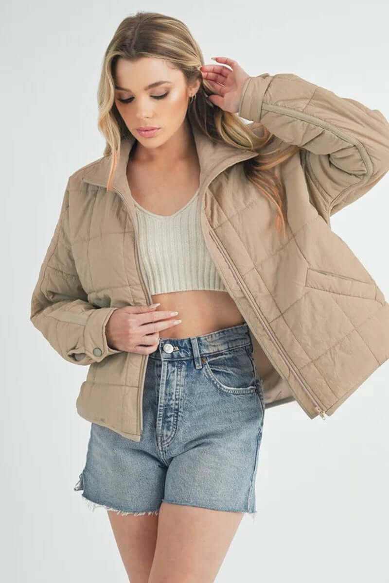 Woman wearing a beige Bebe puffer jacket with oversized silhouette, styled with a crop top and denim shorts.