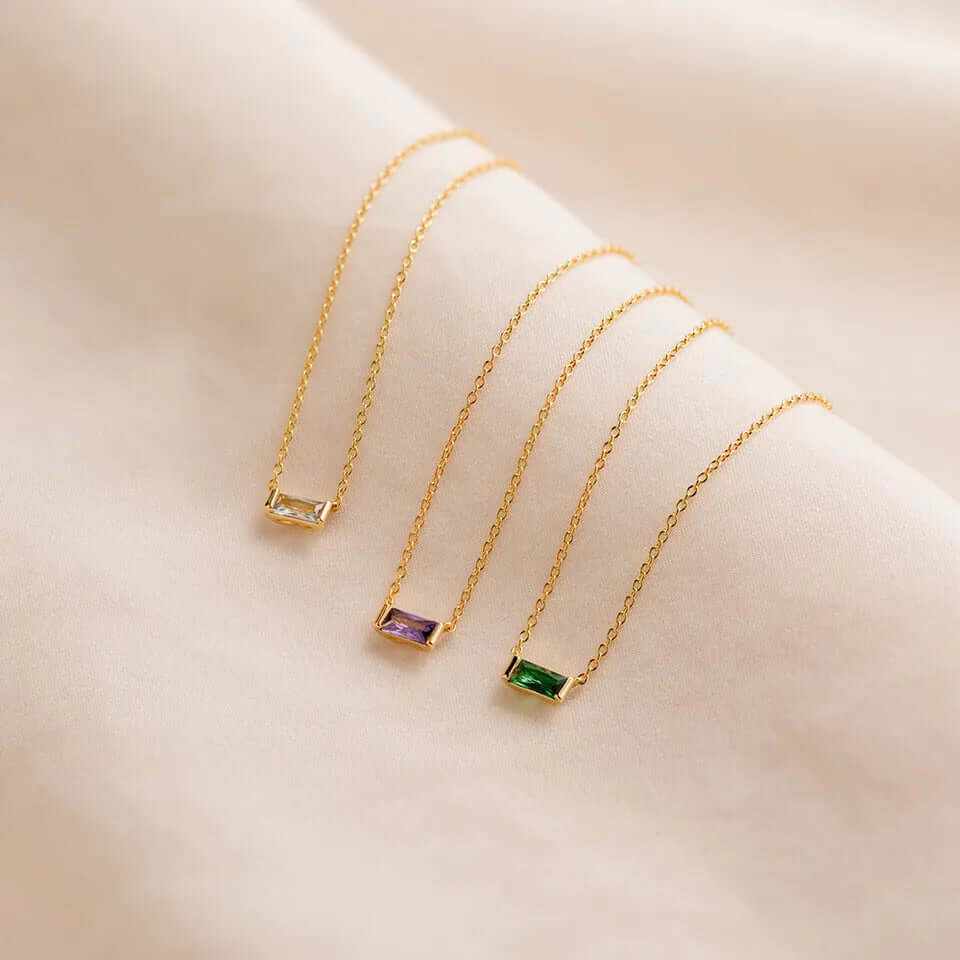 Dainty baguette birthstone CZ charm necklaces on a soft silk background, showcasing vibrant gemstones and gold chains.