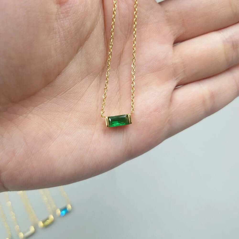 Dainty 18K gold-plated baguette birthstone necklace with green CZ charm, held in hand.
