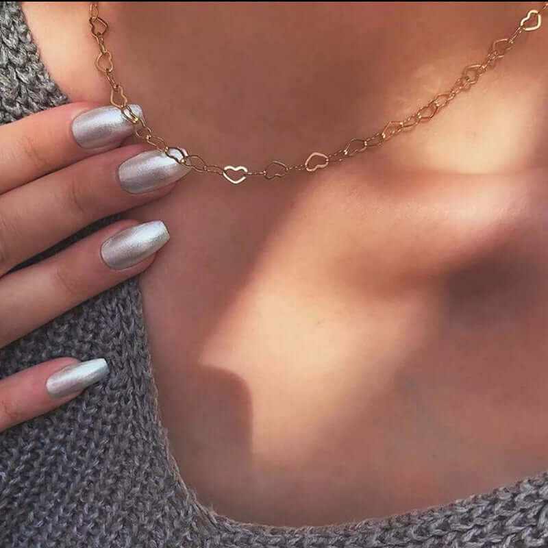 Dainty hollow heart choker necklace in gold plating, worn with silver nails against a cozy sweater.