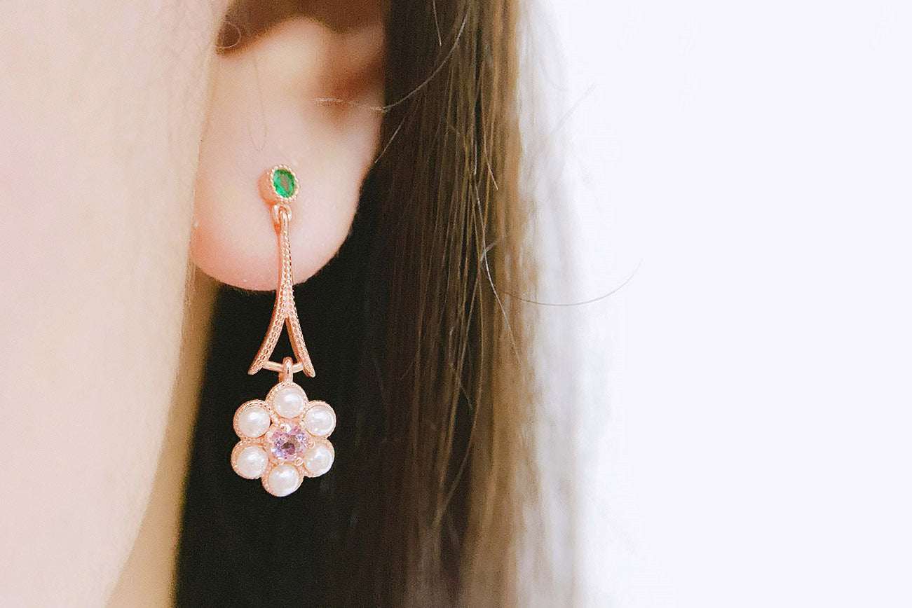 close-up of a woman's ear wearing a delicate flower-shaped earring with pearls and an amethyst center, featuring a small green gemstone stud