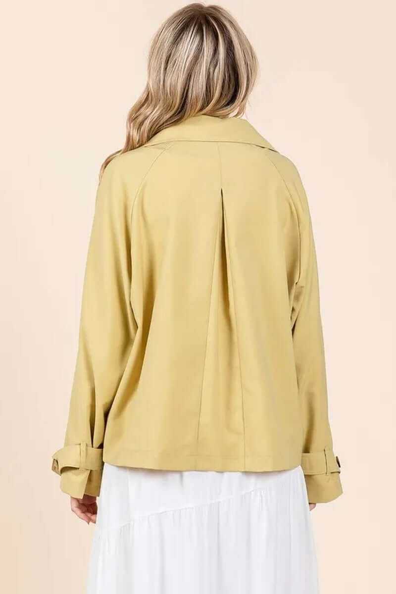 Back view of a woman wearing a yellow double-breasted trench coat jacket over a white dress.