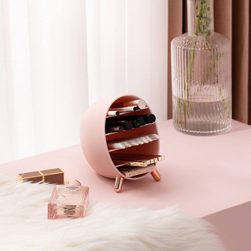 Round pink multi-layer vanity makeup box with compartments on pink table next to perfume bottle and vase