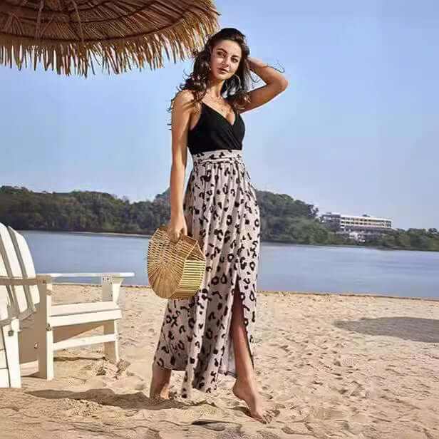 Woman wearing a stylish V-neck wrap spaghetti strap dress with a high waist and leopard print long skirt, standing on the beach carrying a wooden handbag