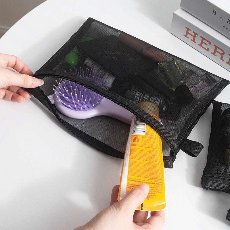 Black transparent mesh makeup organizer bag with zipper holding cosmetic items and hairbrush on white surface