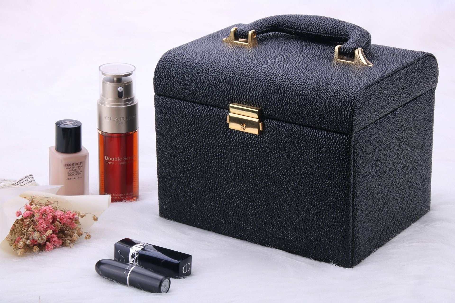 Black water-resistant outdoor makeup case with handle and gold clasp alongside cosmetics and dried flowers