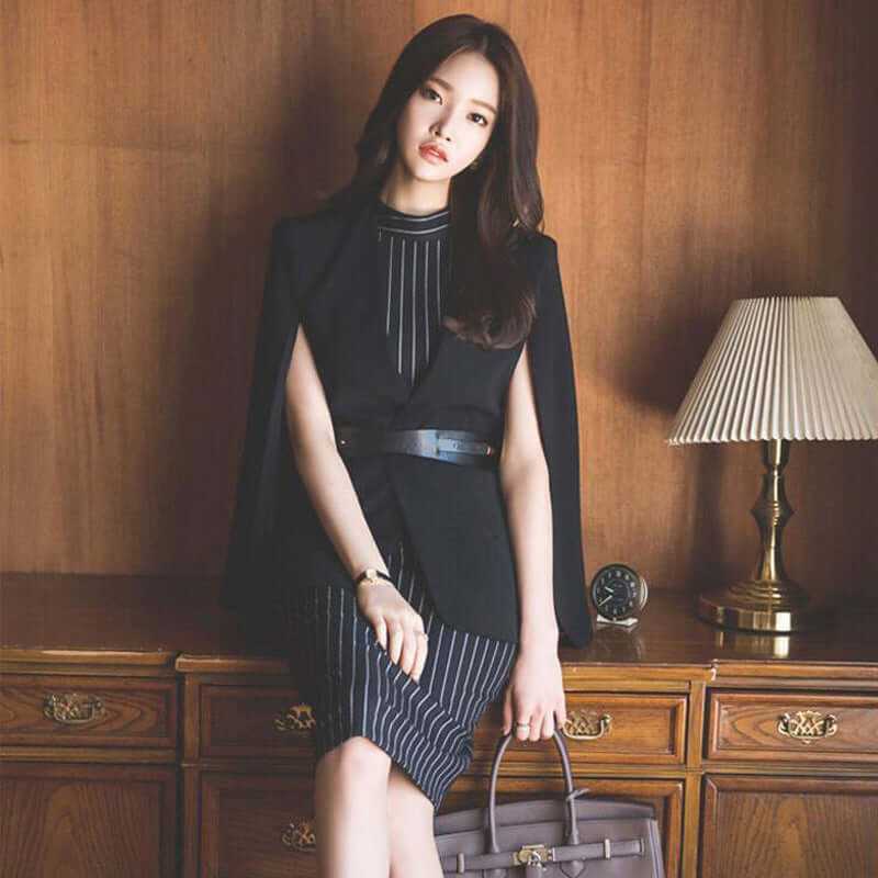 Elegant woman in a black and striped dress with a cape, posing by a vintage wooden dresser and lamp.