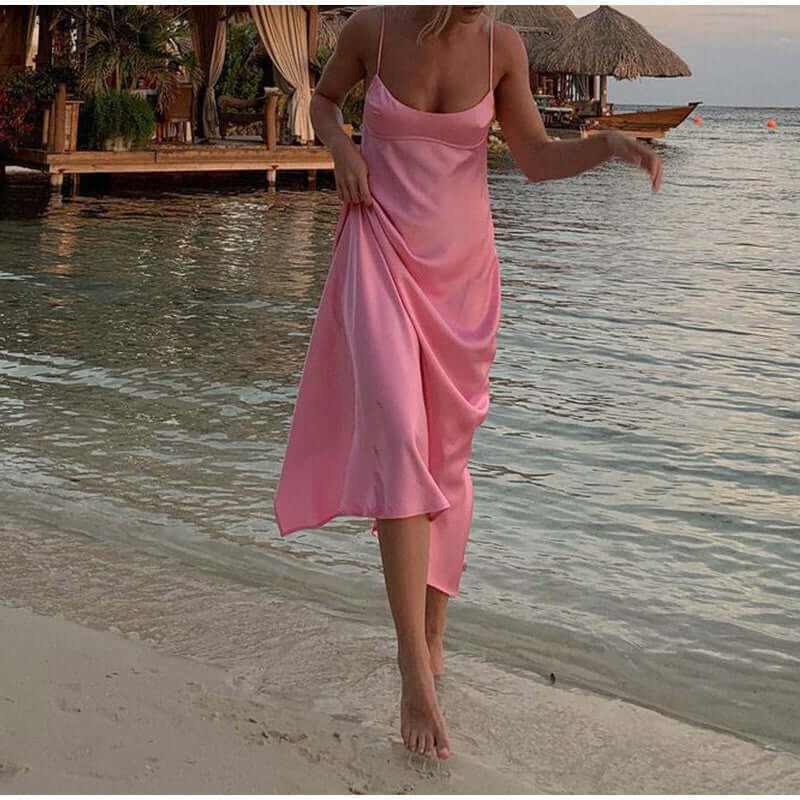 Woman in a pink satin long dress walking by the beach, showcasing a backless design and elegant summer vibe.