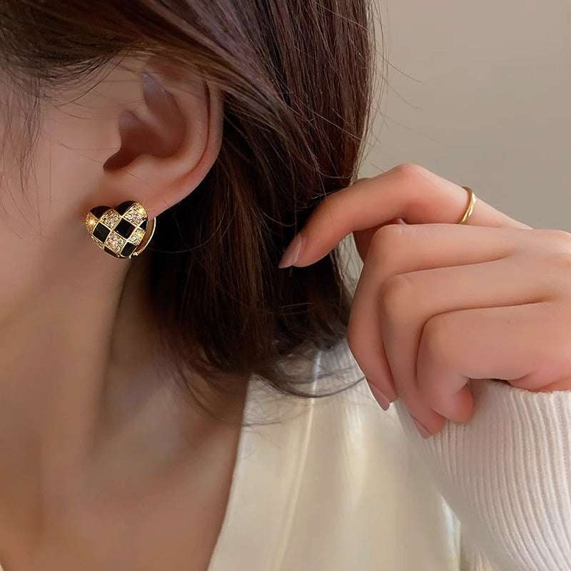Close-up of woman's ear wearing heart-shaped plaid stud earrings in black, gold, and white electroplated copper design