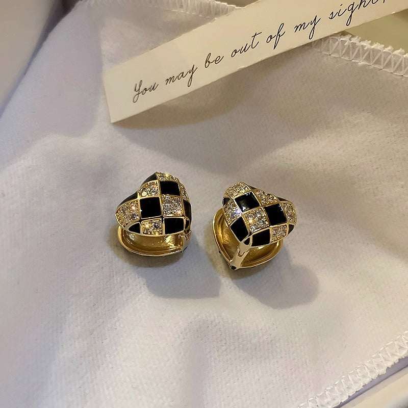 Heart-shaped plaid stud earrings in black and gold with rhinestone accents on white fabric background